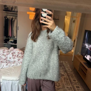 Ljusgrå fluffig stickad tröja - Supermjuk och oversized stickad tröja i ljusgrå ton. Tröjan har lång ärm, rund halsringning och en riktigt fluffig känsla. Perfekt för lager-på-lager och chill dagar. Enkel att matcha med både jeans och mjukisbyxor.