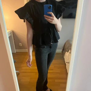 Svart volangblus med vida ärmar - Supersöt svart blus med volanger och vida korta ärmar. Toppen har en lös passform och volangdetalj nedtill. Perfekt att styla med jeans!! 