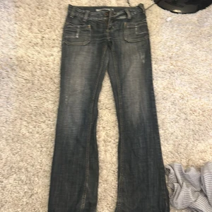 Blå bootcut jeans med fickdetaljer - Säljer ett par blå jeans med bootcut-modell och snygga fickor fram och bak. Jeansen har låg midja och är gjorda i klassiskt denimtyg med slitningar för en cool look. Perfekta för dig som gillar en avslappnad och trendig stil.