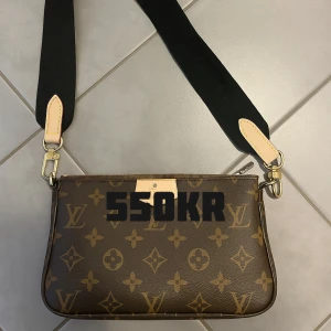  axelrems väska - Snygg axelväska  i klassiskt brunt monogrammönster. Väskan har en bred svart axelrem med detaljer i beige och guldiga metalldelar. Perfekt för dig som vill ha en ikonisk och trendig accessoar.
