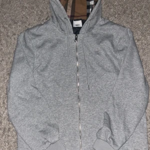Burrbery zip-up Hoodie - Säljer en grå hoodie med dragkedja från Burberry. Jackan har klassiskt rutigt foder i huvan och insidan, samt ribbade muddar. Perfekt för dig som gillar stilrena plagg med exklusiv känsla. Är köpt i storlek S.