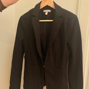 Svart kavaj med klassisk passform - Stilren svart kavaj med klassisk krage och långa ärmar. Kavajen har två fickor framtill och en figurnära passform som ger en snygg siluett. Perfekt att matcha med skjorta eller t-shirt för en clean look.