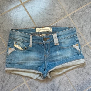 Blå låg midja  jeansshorts från Stradivarius  - Snygga blå jeansshorts från Stradivarius med uppvikta ben och coola detaljer på bakfickorna i vitt och brunt. Klassisk femficksmodell med nitar och bälteshällor. Perfekta för varma dagar och en avslappnad stil.