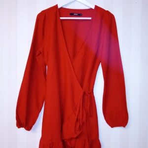 Super fint röda färg omlottklänning med Bik Bok märke, S 38 - Super fint röda färg omlottklänning med Bik Bok märke i kort modell med långa ärmar och knyt i midjan. Klänningen har volangdetalj nertill och är tillverkad i en skön mix av linne och viskos. Perfekt för dig som vill sticka ut med en färgstark look! Kan skickas med extra pris via post.