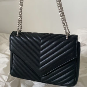 Svart quiltad axelväska Gina Tricot - Snygg svart axelväska från Gina Tricot med quiltad design och silvrig kedjerem. Väskan har ett klassiskt kuvertformat lock och stängs med magnetknapp. Perfekt storlek för det viktigaste och ger en trendig vibe till din outfit.