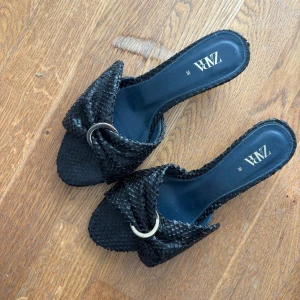 Svarta sandaler med struktur från Zara - Snygga svarta sandaler från Zara med strukturerad yta som påminner om ormskinn. De har öppen tå, platt sula och en dekorativ guldfärgad ring framtill. Perfekta för att lyfta en somrig outfit. Obs klister på undersidan 
