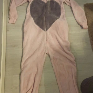 Rosa fleece onesie med hjärta & huva - Supermysig rosa fleece onesie från Love to Lounge i storlek S. Har ett stort grått hjärta på magen, dragkedja framtill, huva med roliga ansiktsdetaljer och öron samt en svans baktill. Perfekt för chilliga dagar hemma eller temakvällar!