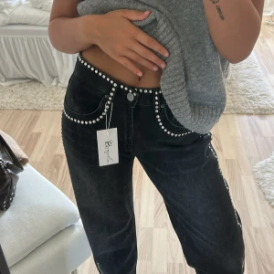 Svarta jeans med nitar från Design by Si XXS - Svarta jeans med coola silvriga nitar längs fickor och sidor. Modellen är rak och har en relaxed fit med låg midja. Jeansen har klassisk femficksdesign och är tillverkade i denim. Perfekta för dig som vill sticka ut med detaljer. Sitter tajt vid midjan och långa vid fötter, är 160. 
