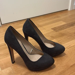 Svarta mocka pumps med hög klack - Stilrena svarta pumps från Tamaris i mjuk mocka. Skorna har hög klack och plattform framtill, samt en klassisk spetsig tå. Perfekta för dig som vill ha en elegant och tidlös look med extra höjd.