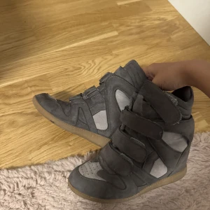 Gråa wedge sneakers med kardborre  - Snygga grå sneakers med inbyggd kilklack och coola paneler i mocka och textil. Skorna har flera breda kardborreband och en platt gummisula. Perfekta för dig som vill ha lite extra höjd men ändå en sportig vibe.