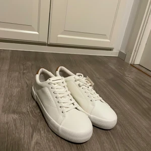 Vita sneakers från Bershka strl 41 - Snygga vita sneakers från Bershka i storlek 41. Skorna har en stilren design med vita snören, platt sula och bruna detaljer vid hälen. Tillverkade i syntetmaterial med klassisk rund tå. Perfekta för dig som gillar en clean och fräsch look.