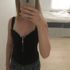 Snygg svart body från River Island - Svart korsettopp från River Island med breda axelband och snygg silverfärgad dragkedja framtill. Toppen har volangkant och figurnära passform. Perfekt att styla med jeans eller kjol för en cool look.