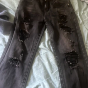 Svarta ripped jeans med dragkedja - Svarta jeans med slitna och trasiga detaljer längs benen för en edgy look. Klassisk femficksmodell med dragkedja och knapp framtill. Insidan av linningen har ett färgglatt mönster. Perfekta för dig som gillar streetstyle.