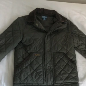 Quiltad grön jacka från Polo Ralph Lauren - Snygg quiltad jacka från Polo Ralph Lauren i mörkgrönt med bruna detaljer och krage i manchester. Klassisk look med tryckknappar och två fickor framtill. Perfekt för dig som vill ha en stilren och tidlös jacka med skön passform.PRIS kan diskuteras vid snabb affär 