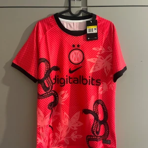 Inter Milan Nike fotbollströja röd - Inter Milan fotbollströja i röd med svarta detaljer och tryck. Tröjan har korta ärmar, svart krage och är tillverkad i lätt, ventilerande material. Snyggt mönster med ormar och blommor samt klubbmärke och sponsortryck på bröstet.