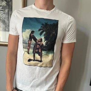 Vit Limitato t-shirt, Riocam - Vit limitato t-shirt med stor färgglad grafisk print framtill som föreställer två personer vid en pool och palmer i bakgrunden. T-shirten har rund halsringning och korta ärmar. Materialet känns mjukt och lät. Skick 7/10, storlek M. Title; Who is. Finns box 