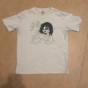 Vit t-shirt med grafiskt tryck - Vit t-shirt från HUF i storlek L med ett stort grafiskt tryck på framsidan som visar två tecknade händer och gröna siffror. Klassisk passform och rund halsringning. Perfekt för dig som gillar streetwear och vill sticka ut lite extra.