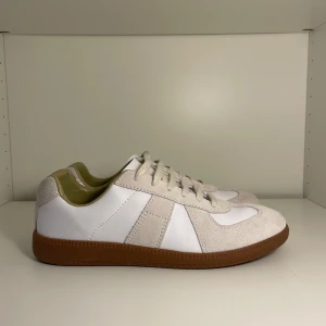 Maison margiela vit/beige -  sneakers i vitt skinn och beige mocka med klassisk gummisula. Snygg retrostil med rund tå och snörning. Perfekta för dig som gillar minimalistisk och stilren design.