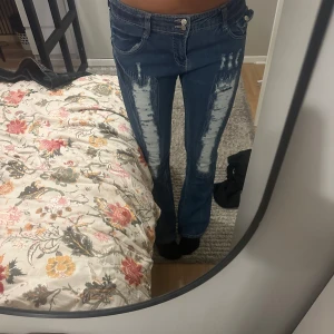Blå bootcut jeans med slitningar - Säljer ett par blå bootcut jeans med kraftiga slitningar framtill på båda benen. Jeansen har låg midja, klassiska fickor och en snygg tvättad look. Materialet är jeans och passformen är normal med lite utsvängda ben. !Kolla gärna in mina andra annonser!