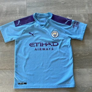 Manchester City Puma matchtröja blå 2019 hemma - Manchester Citys officiella matchtröja från Puma  i ljusblått med mörkblå detaljer på axlarna. Broderad klubbmärke och Puma-logga, samt tryckt sponsor på bröstet. Tillverkad i ventilerande polyester för maximal komfort på planen.