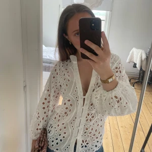 Vit broderad blus med knappar - Superfin vit blus i bomull med broderade blommor och hålmönster. Djup v-ringning, knappar framtill och vida ärmar med resår vid handlederna. Perfekt till jeans för en fräsch och somrig look. Har en liten fläck men tror den går bort i tvätten