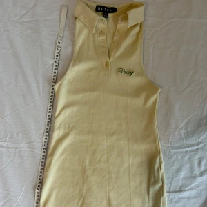 Gul ärmlös klänning från Motel - Snygg ljusgul ärmlös klänning från Motel med krage, knappar framtill och broderad grön text 'Vacay' på bröstet. Klänningen är i mjuk bomull och har en sportig, tight passform som känns både chill och trendig.