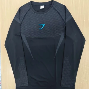 Svart långärmad träningströja Gymshark - Svart långärmad träningströja från Gymshark med gråa geometriska detaljer och blå logga på bröstet. Det är gymshark onyx v5 kollektionen och den säljs ej längre. Helt ny.
