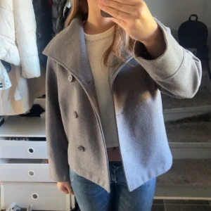 Grå rock med bred krage - Snygg grå rock med bred krage och dubbelknäppning framtill. Jackan har en rak, lite kortare passform och är perfekt för dig som gillar stilrena och klassiska plagg. Passar till jeans eller dressat, och är lätt att matcha med olika outfits.