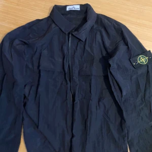 Svart overshirt från Stone Island - Säljer en svart overshirt från Stone Island med klassisk patch på ärmen. Jackan har dragkedja framtill, flera fickor och är tillverkad i lätt polyester. Perfekt för lager-på-lager och streetwear-vibe. Passar perfekt nu i sommar då materialet är tunt som en windbreaker