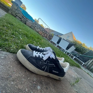 Premiata svarta sneakers med vit sula - Snygga svarta sneakers från Premiata med vit sula och vita skosnören. Skorna har en tydlig logga på sidan och är tillverkade i mocka med detaljer i skinn. Perfekta för dig som gillar en clean och stilren look.