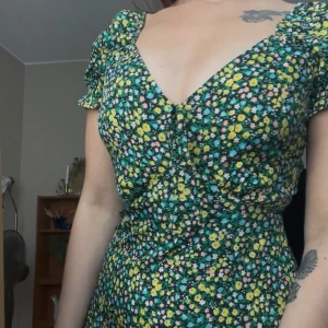 Blommig kortklänning med v-ringning - Supersöt kortklänning med blommigt mönster i grönt, gult, vitt och rosa. Klänningen har v-ringning, puffiga korta ärmar med volangkant och markerad midja. Perfekt för sommarens alla tillfällen!