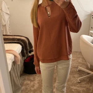 Rostfärgad blus med v-ringning - Säljer en snygg rostfärgad blus med v-ringning och dekorativ detalj vid halsen. Blusen har långa ärmar med knapp vid ärmslut och en avslappnad passform. Perfekt att styla med jeans eller kjol för en trendig look.