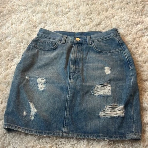 Blå jeanskjol med slitningar från Monki - Säljer en blå jeanskjol från Monki i storlek XS. Kjolen är kort och har snygga slitningar framtill för en cool, avslappnad vibe. Klassisk femficksmodell med knapp och dragkedja framtill. Perfekt till sneakers eller boots. 