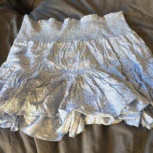 Blommig blå volangkjol från 157 - Supersöt ljusblå kortkjol från 157 med små vita blommor och volangdetaljer. Kjolen har smock i midjan för en bekväm passform och är perfekt för varma dagar. Lätt och luftig känsla, passar dig som gillar en feminin och trendig stil.