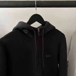 Svart hoodie från Hugo Boss - Svart hoodie från Hugo Boss med dragkedja framtill och diskret logga på bröstet. Tröjan har huva med snörning och är tillverkad i mjukt bomullsmaterial. Perfekt för en clean och stilren look.