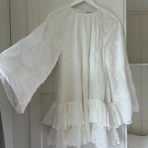 Vit blommig volangklänning från NA-KD - Superfin vit kortklänning från NA-KD med stora blommiga broderier, volanger nertill och vida långärmar. Klänningen har en luftig passform och är perfekt för sommarens alla tillfällen. Tunn och lätt i materialet, riktigt söt och trendig.