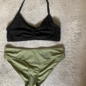 Svart och grön bikini med enkel design - Snygg bikini med svart topp och olivgröna trosor. Toppen har smala axelband och lätt rynk framtill för en stilren look. Trosorna är högt skurna och har en enkel, klassisk passform. Perfekt för strandhäng eller poolparty.