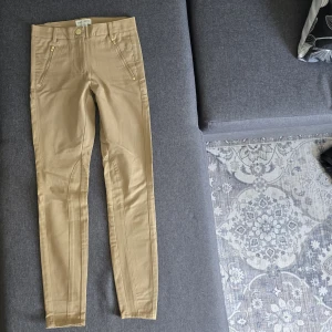 Beiga skinny byxor Holly & Whyte - Snygga beiga byxor från Holly & Whyte med skinny passform och guldfärgade dragkedjor framtill. Byxorna har midja med hällor, dekorativa sömmar och är tillverkade i bomullsmix. Har aldrig använd.