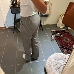 Grå randiga kostymbyxor - Snygga grå kostymbyxor med vita vertikala ränder och raka ben. Byxorna har hög midja och klassisk passform, perfekta för en clean och stilren look. Materialet känns mjukt och följsamt, och de har fickor i sidorna.