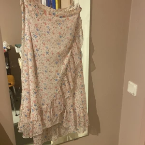 Blommig omlottkjol med volang - Supersöt långkjol i ljusbeige med små blommor i rosa, blått och rött. Kjolen har omlottmodell och volangkant som ger en romantisk vibe. Perfekt för vår och sommar när du vill känna dig extra fin.