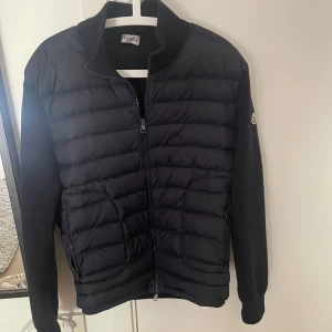 Svart Moncler pufferjacka herr - Svart pufferjacka från Moncler med quiltad front och släta ärmar. Jackan har dragkedja framtill, ståkrage och diskret Moncler-logga på ärmen. Perfekt för dig som vill ha en stilren och exklusiv look under höst och vinter.