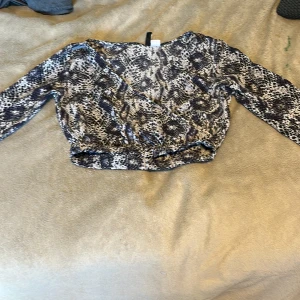 Leopardmönstrad omlottopp från H&M - Säljer en croppad omlottopp från H&M Divided i storlek 32. Toppen har ett coolt leopardmönster i svart, grått och vitt, långa ärmar och resår i midjan för en snygg passform. Perfekt för dig som vill sticka ut med en trendig look!