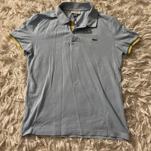 Ljusblå pikétröja från Lacoste - Snygg ljusblå pikétröja från Lacoste med klassisk krage och knappslå. Tröjan har gula detaljer vid ärmslut och krage, samt den ikoniska krokodilloggan på bröstet. Perfekt för en stilren look!