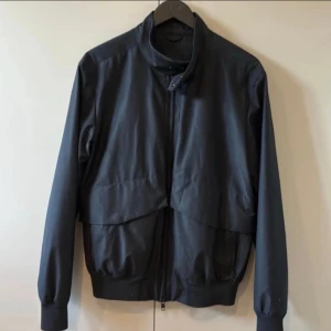 Svart jacka från J.Lindeberg - Snygg svart jacka från J.Lindeberg tillverkad med Loro Piana Storm System-tyg. Jackan har en stilren design med dragkedja och långa ärmar. Perfekt för en modern och elegant look.