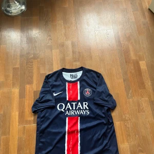 Blå PSG fotbollströja - Säljer en blå PSG fotbollströja från Nike med röd och vit rand på framsidan. Tröjan har korta ärmar och är tillverkad i Dri-FIT material för optimal komfort. Perfekt för fotbollsfans!