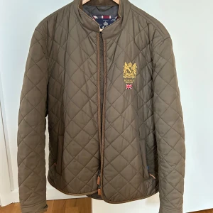 Brun quiltad jacka från Morris - Säljer en stilren brun quiltad jacka från Morris med broderad logga på bröstet. Jackan har en dragkedja framtill och en klassisk krage. Perfekt för höst och vår. Nypris 2199 kr