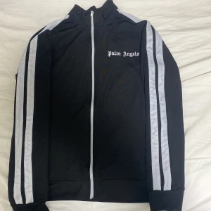 Svart träningsjacka från Palm Angels - Snygg svart träningsjacka från Palm Angels med vita ränder längs ärmarna och dragkedja framtill. Jackan har en broderad logga på bröstet och är perfekt för en sportig look.