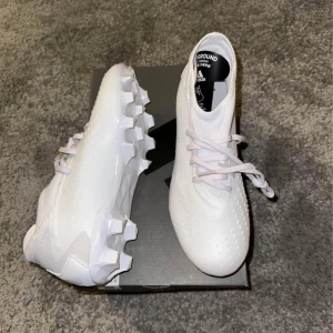Vita fotbollsskor från Adidas - Säljer ett par stilrena vita fotbollsskor från Adidas. Skorna har en modern design med snörning och är perfekta för spel på olika underlag. De har en greppvänlig sula för optimal prestanda på planen.