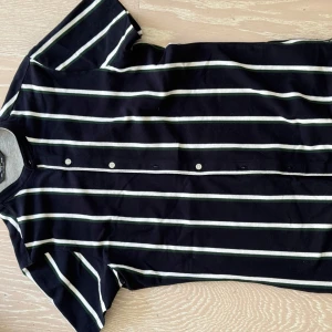 Svart och vit randig t-shirt från Bershka - Säljer en stilren svart t-shirt från Bershka med vita och gröna ränder. T-shirten har korta ärmar och knappar framtill. Perfekt för en avslappnad look.