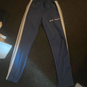 Blå  Palm Angels byxor  - Snygga blå byxor från Palm Angels med vita ränder längs sidorna. Byxorna har en elastisk midja och en avslappnad passform. Perfekta för en sportig look. Äkta såklart bara höra av sig vid frågor passar även s 
